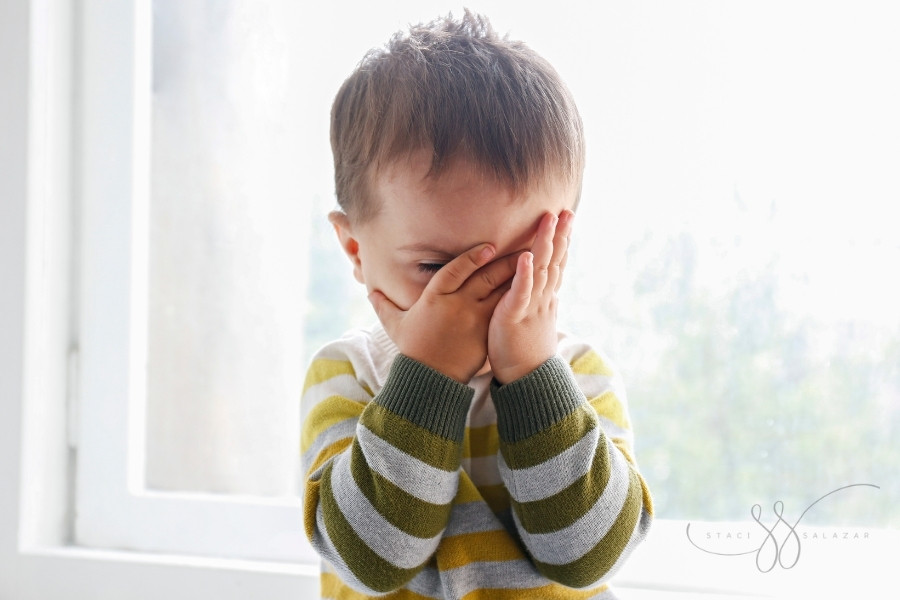 upset toddler covering their face sitting on a windowsill