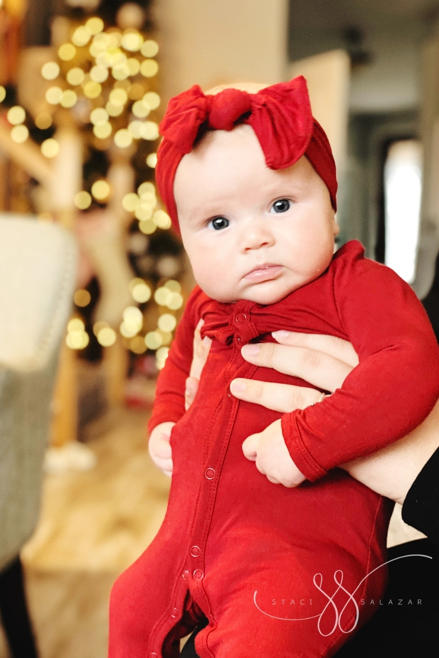 baby in red sleeper and bow looking at camera with bokeh holiday lights in background