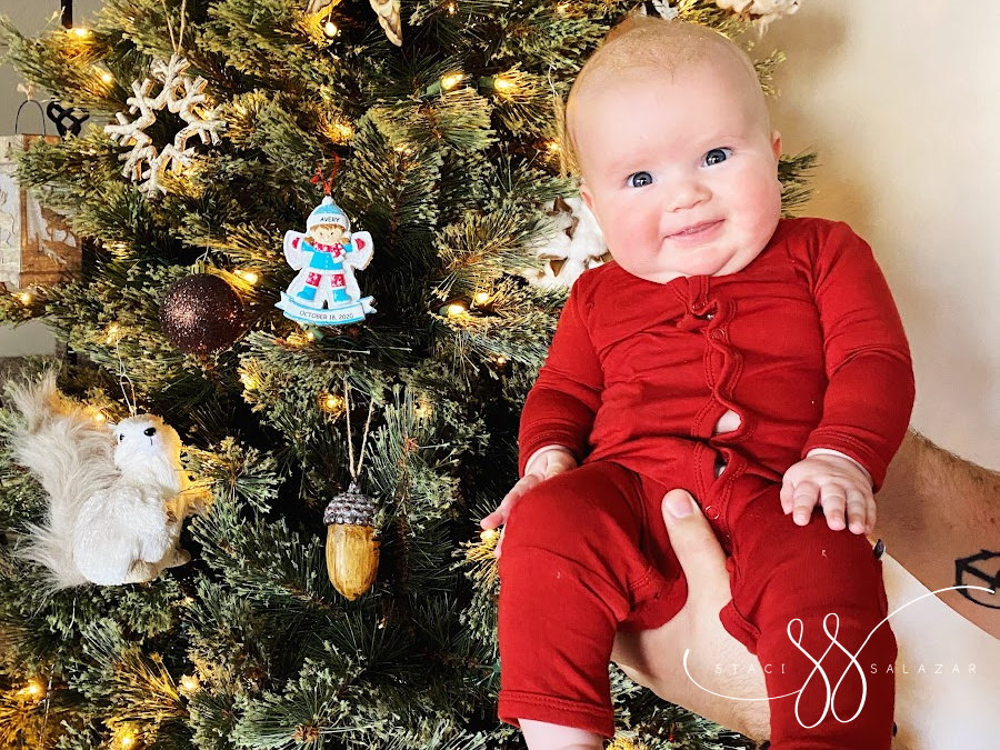 baby in red sleeper in front of Christmas tree with personalized ornament