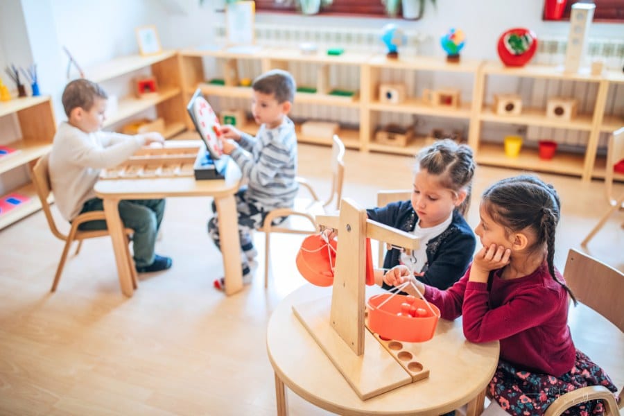 Two preschool age girls sitting together in their classroom and playing with scale. There are other school boys playing in the background.