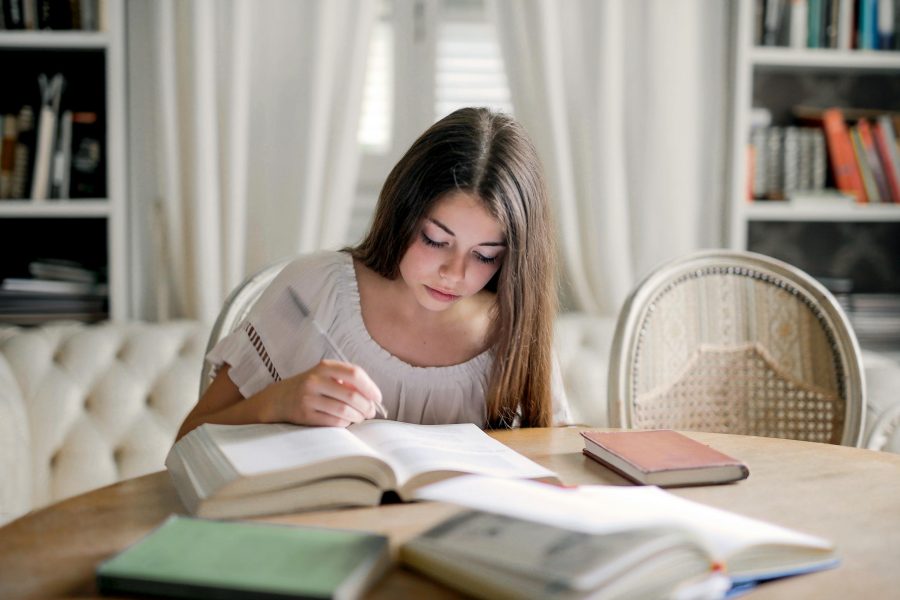 A young girl focuses on studying at a table, surrounded by books, indoors.